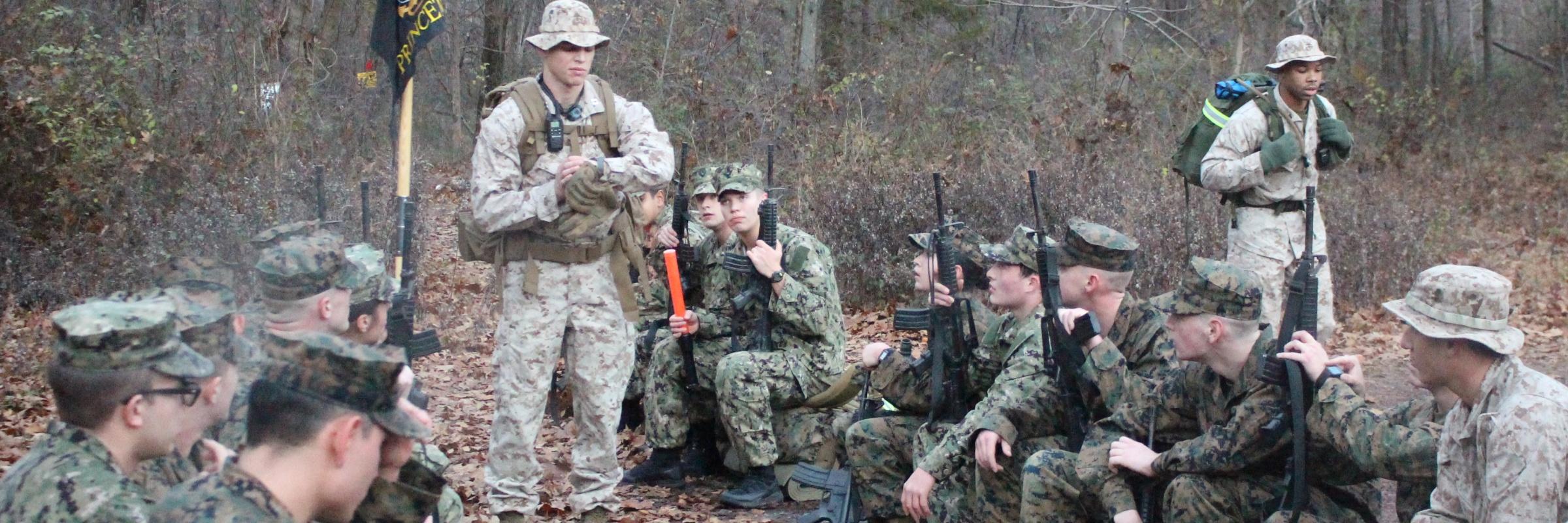 A group of midshipmen in the woods with rifles are briefed by Captain Paragone on a Marine Field Exercise.