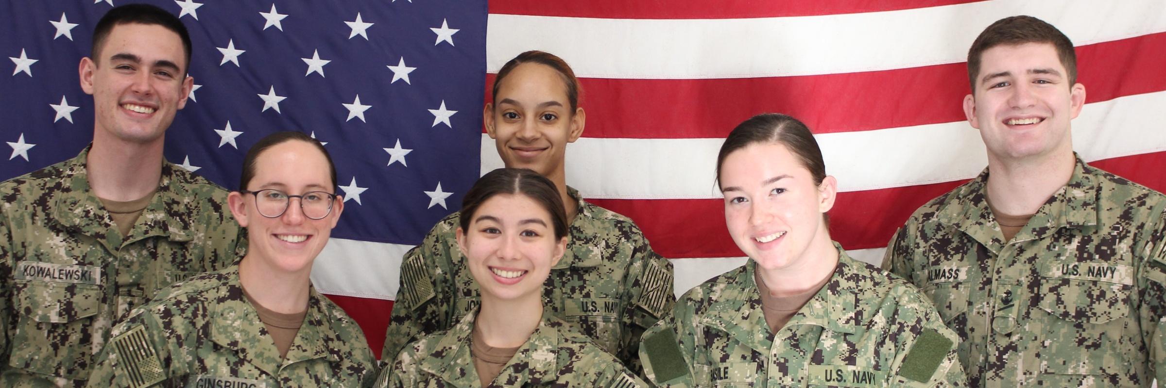 Six smiling midshipmen are standing in front of an American Flag.