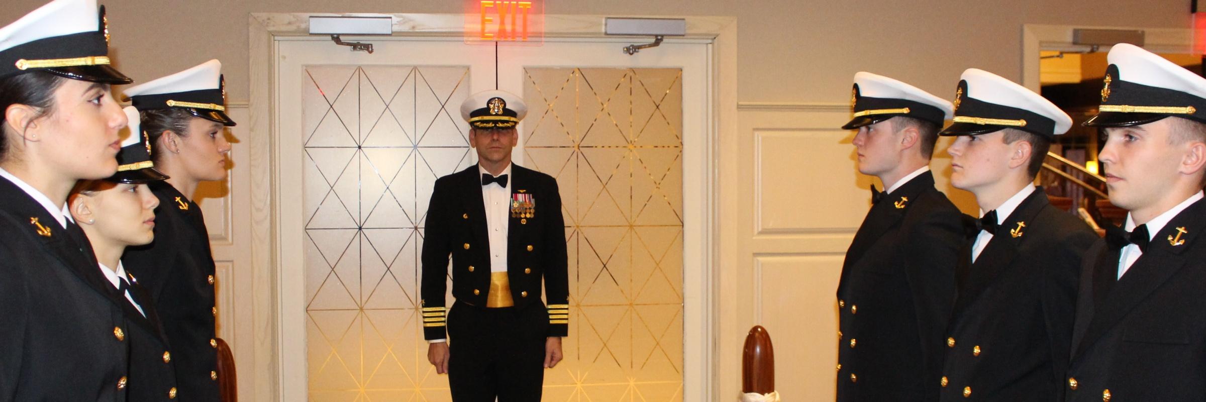 Captain Paul De Marcellus prepares to make his official entry into the military ball, with a group of midshipmen in dress uniforms standing at the ready to salute upon entry.