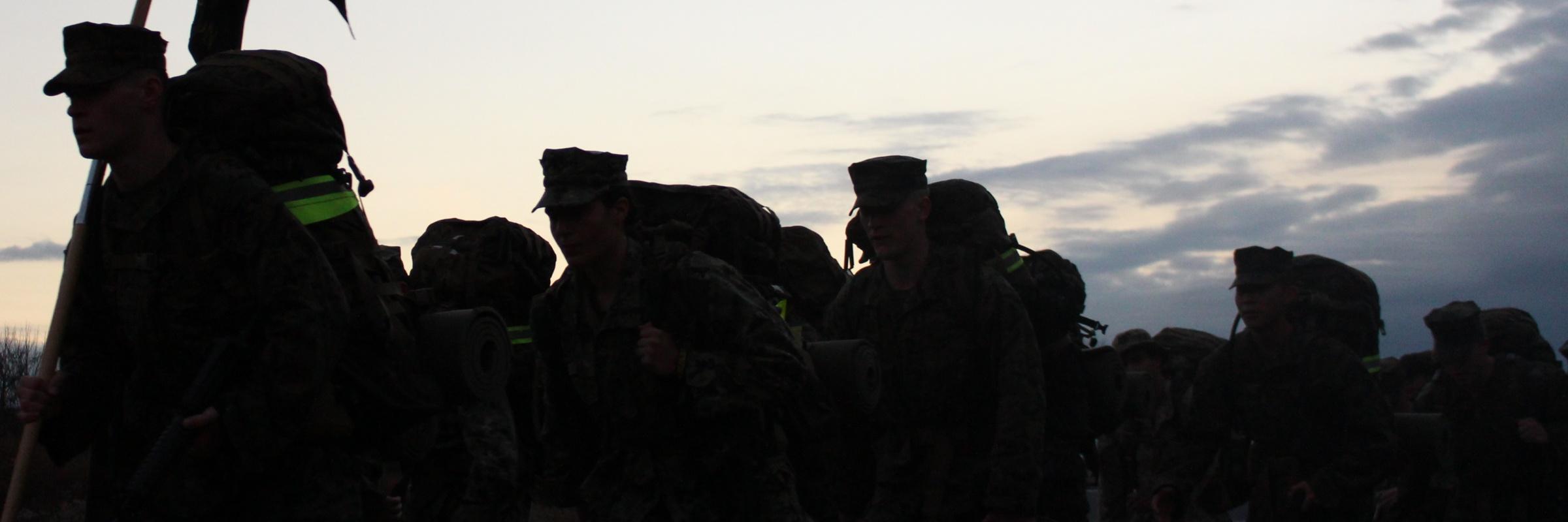 A group of midshipmen with weighted packs, on a hike, are silhouetted in front of the dawn sky.